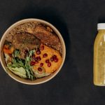 A vibrant vegan bowl with quinoa, fresh vegetables, and a smoothie bottle against a dark backdrop.